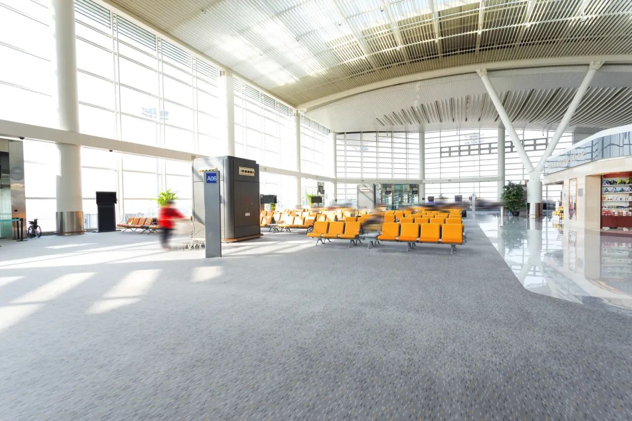 A bright airport terminal hall with a glass wall, grey carpet, rows of orange seats and a single traveler walking past, the kind of setting airport facility cleaning services programs are built around