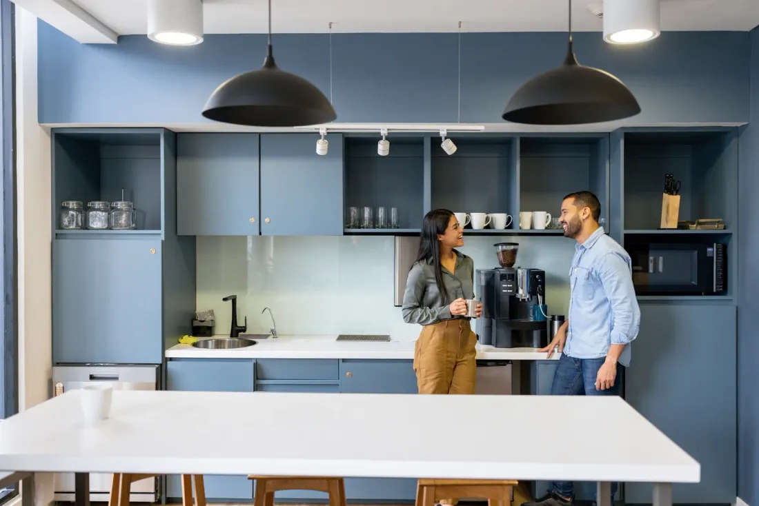 Two people talking by a coffee machine in an office kitchen with slate-blue cabinets, black pendant lamps and a white island, accompanying "Office Cleaning Checklist: Daily, Weekly and Monthly Tasks"