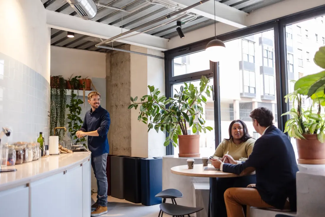 Three people in an office kitchen with plants, one at the counter and two talking at a round table by tall windows, like the offices EBS cleans for tenants in Poway, CA