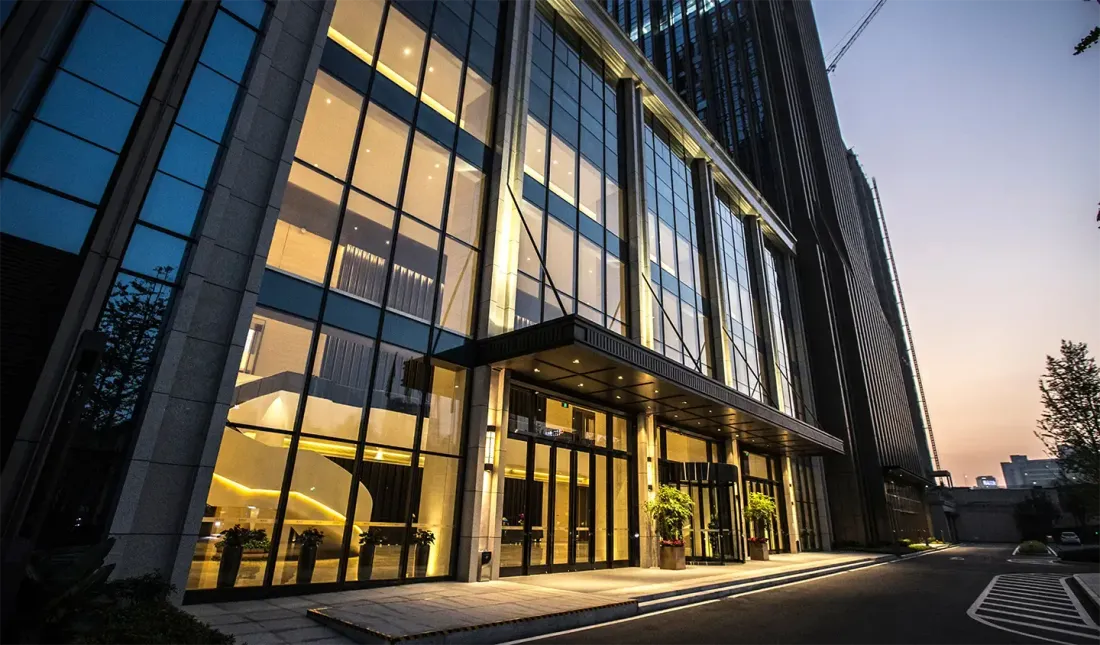A glass-fronted office building lit from inside at dusk, with a canopy over the entrance and potted plants beside the doors, for the EBS guide "Can You Strip and Wax Floors Without Closing the Building?"