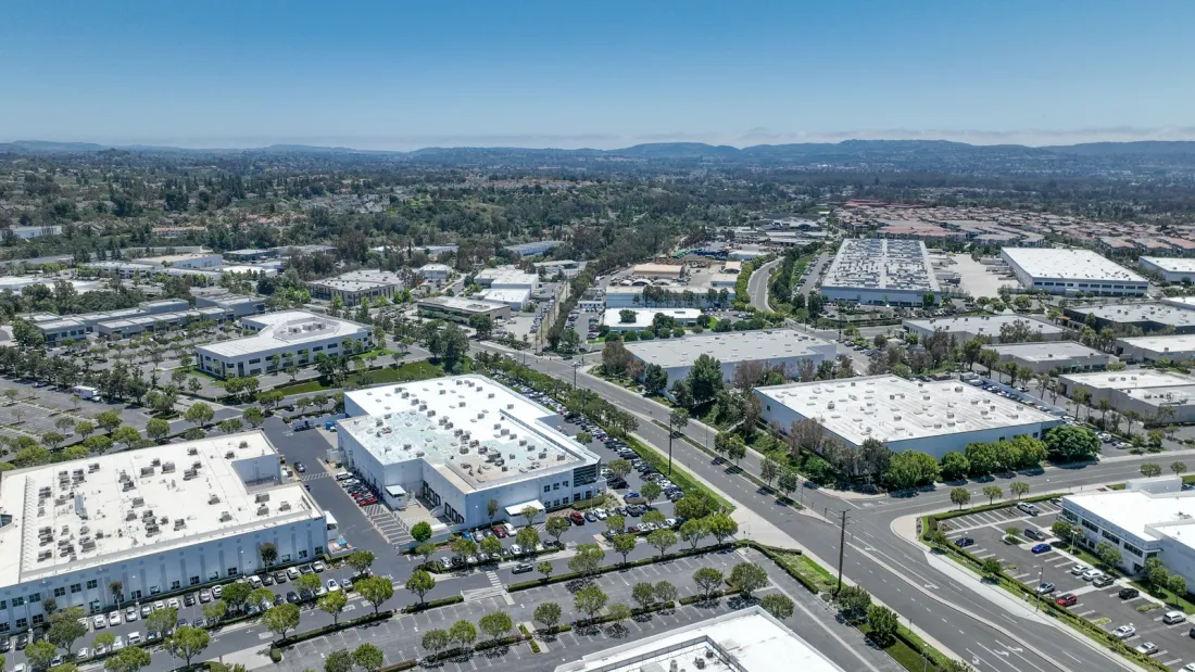 An aerial view of a business park with flat-roofed office and industrial buildings, parking lots and tree-lined streets, with hills on the horizon