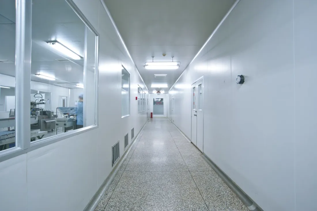 A white corridor with a speckled floor and windows looking into a production room where a gowned worker stands at equipment, for the EBS guide "How Often Should a Data Center Subfloor Be Cleaned?"