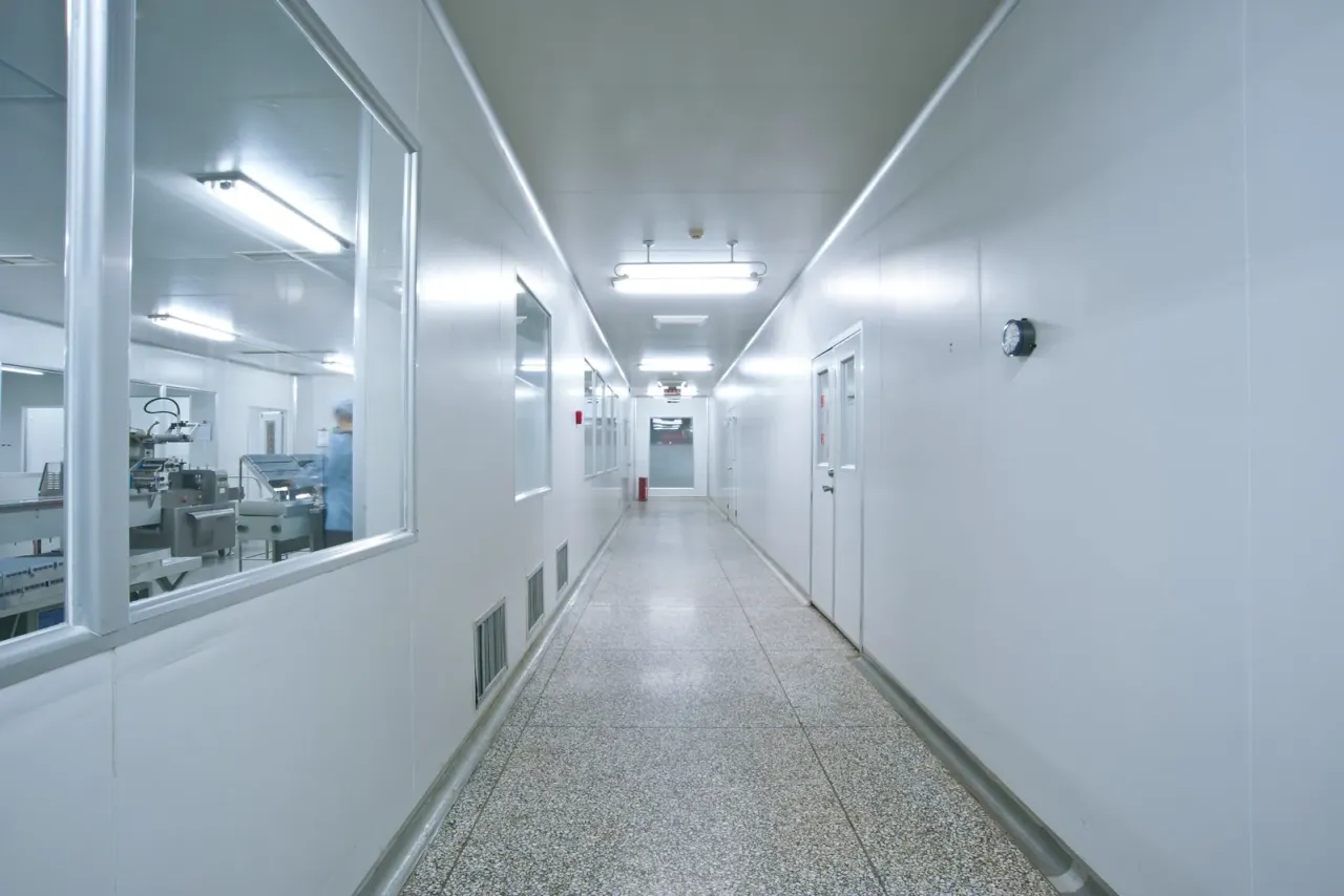 A white corridor with a speckled floor and windows looking into a production room where a gowned worker stands at equipment, an example of the spaces EBS cleans for this sector in Orange County