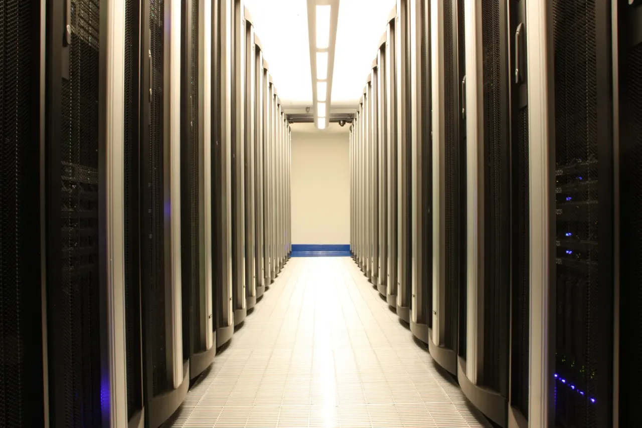 A narrow aisle between rows of black server racks leading to a lit end wall, accompanying "How Often Should a Data Center Subfloor Be Cleaned?"
