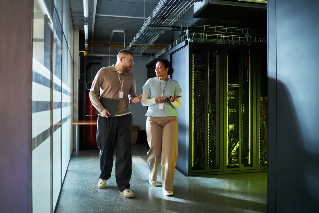 Two people with lanyards walking and talking beside server cabinets in a data center corridor, typical of the facilities covered by data center cleaning in Los Angeles County
