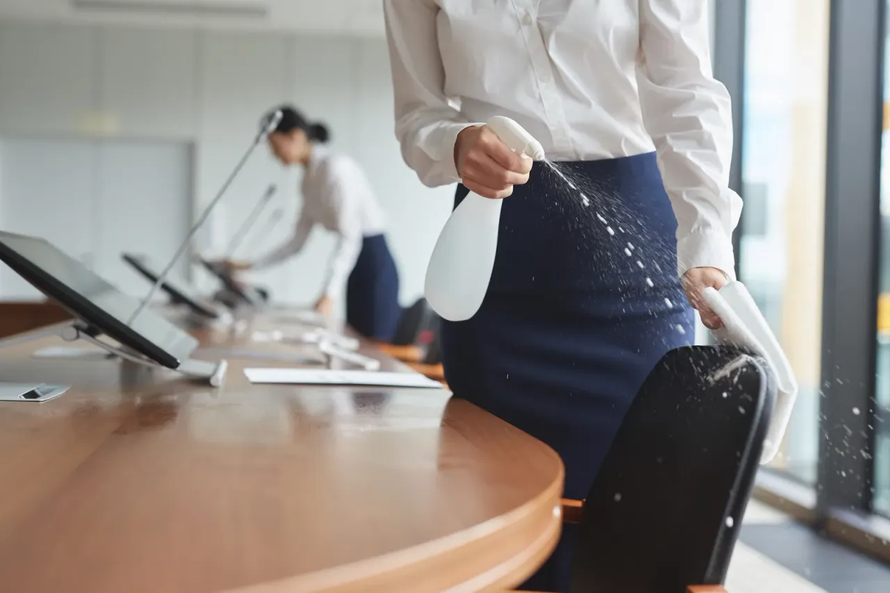 Two women in white blouses spraying and wiping a curved wooden desk with monitors in an office, accompanying "How Much Does Electrostatic Disinfection Cost?"