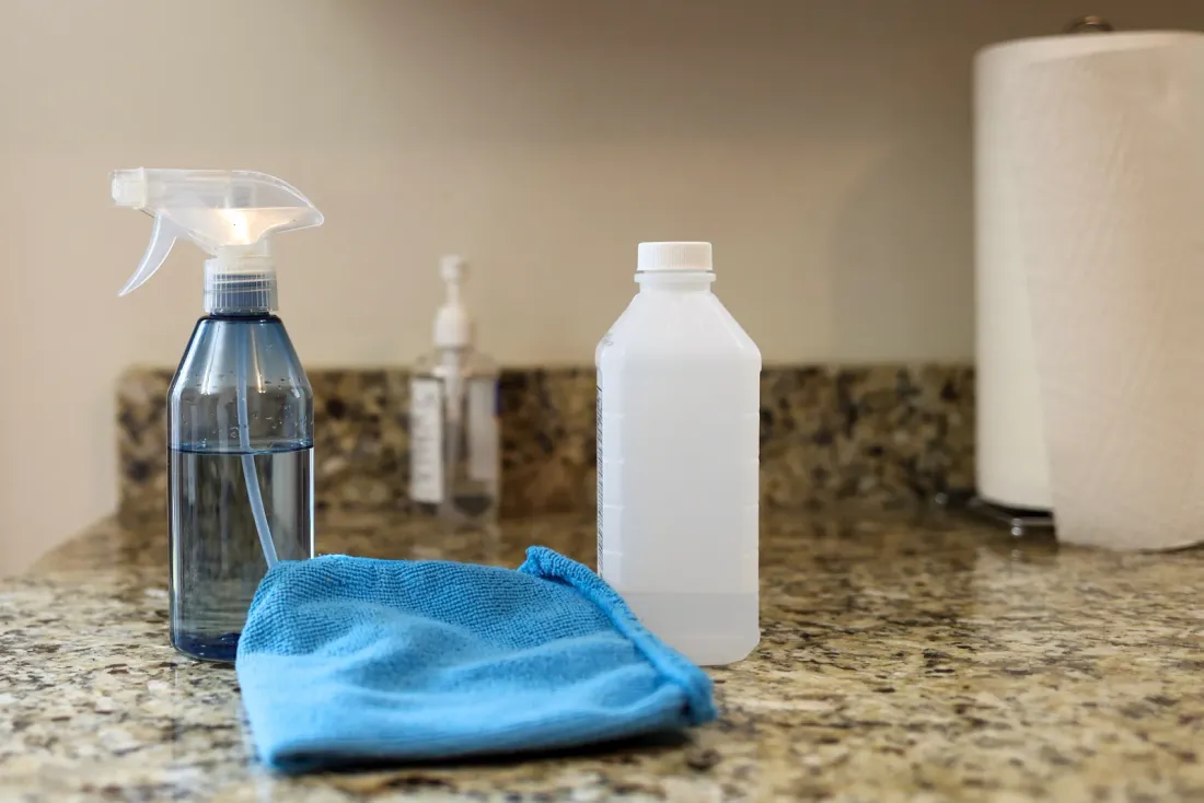 A spray bottle, a plastic bottle, a hand-sanitizer pump and a blue microfiber cloth on a granite counter beside a paper towel roll, for a page on medical office cleaning in Temecula, CA