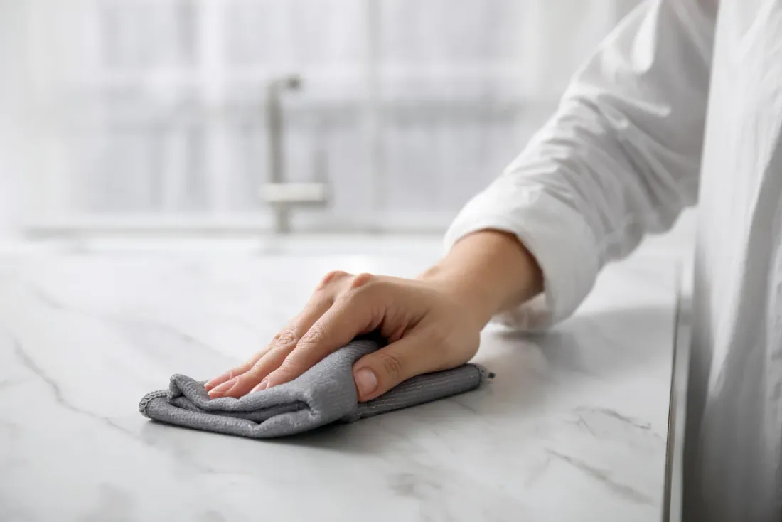 A bare hand wiping a white marble countertop with a grey microfiber cloth, accompanying "How Much Does Cleanroom Cleaning Cost?"