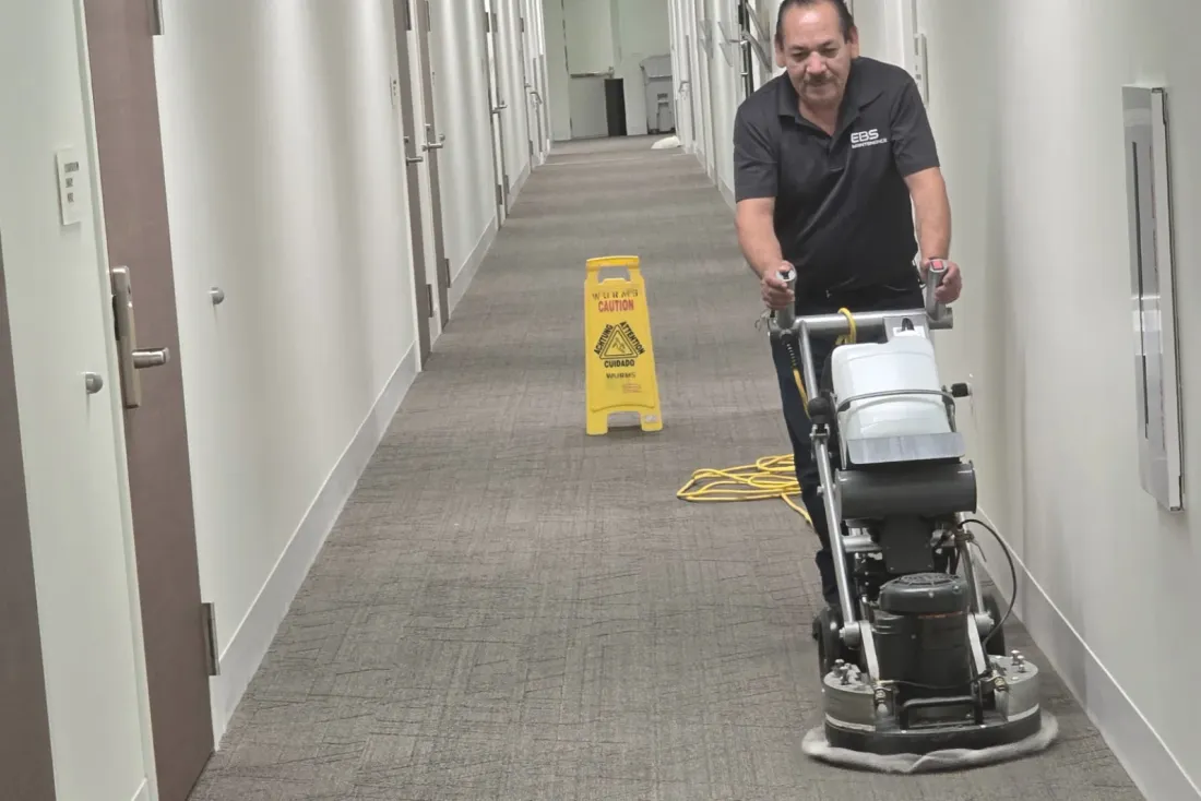 A man in a black polo shirt running a carpet cleaning machine down a carpeted corridor lined with doors, with a yellow caution sign and a power cord behind him, the EBS team behind cleaning programs in Perris, CA