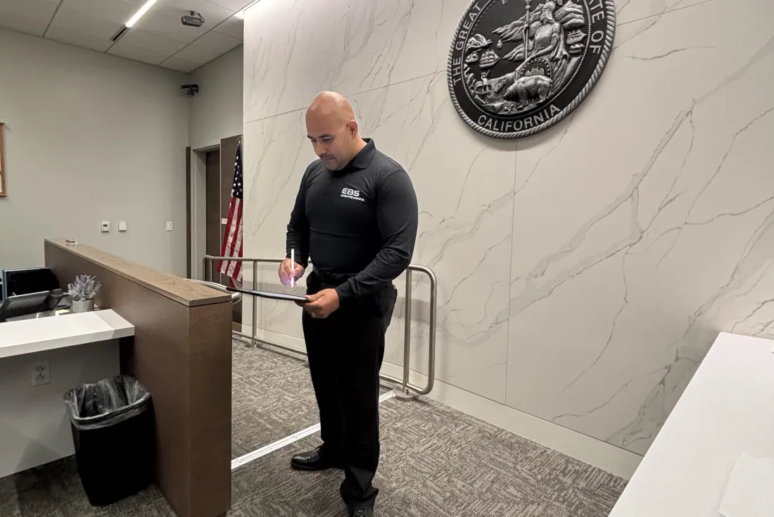 A man in a black shirt with the EBS logo writing on a clipboard in a lobby with a marble wall, a US flag and a large state seal, the EBS team behind cleaning programs in Monrovia, CA