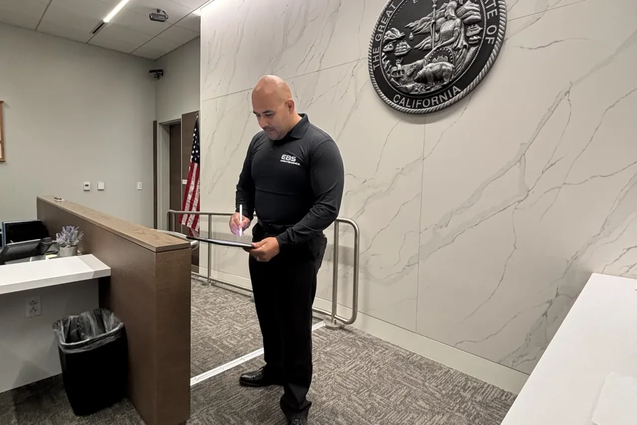 A man in a black shirt with the EBS logo writing on a clipboard in a lobby with a marble wall, a US flag and a large state seal, EBS staff at work, the same crews that serve Vista, CA