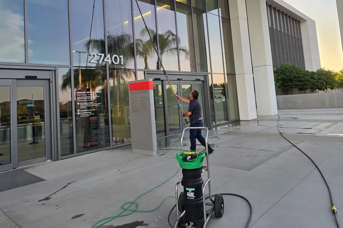 A worker cleaning the glass entrance of a building with a water-fed pole, with a green-topped water cart and hose in the foreground, part of the EBS Maintenance team serving Encinitas, CA