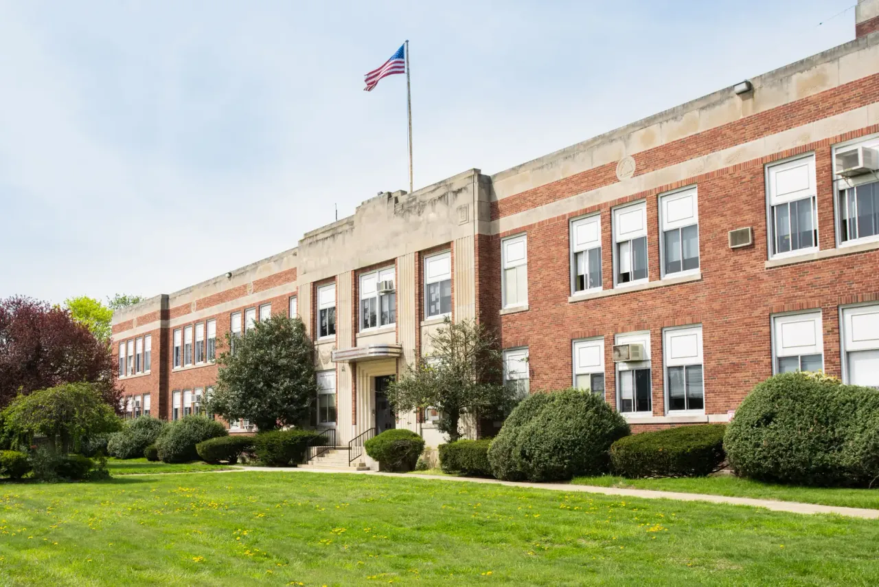 A three-storey red-brick school building with a US flag, a green lawn and trimmed shrubs, illustrating the EBS article "How Many Custodians Does a School Need?"