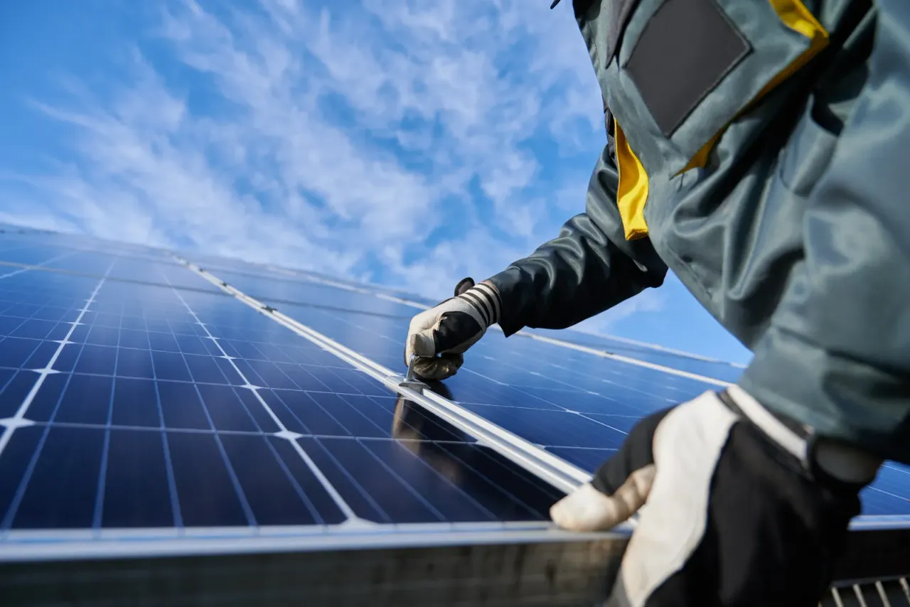A worker in a grey jacket and work gloves handling a solar panel against a blue sky, typical of the facilities covered by energy and utility facility cleaning in Orange County