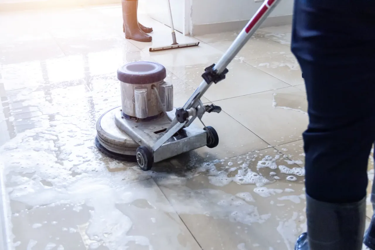 A worker running a rotary floor machine over wet, soapy tile while another person works a floor squeegee in the background, illustrating floor and carpet care in Orange County