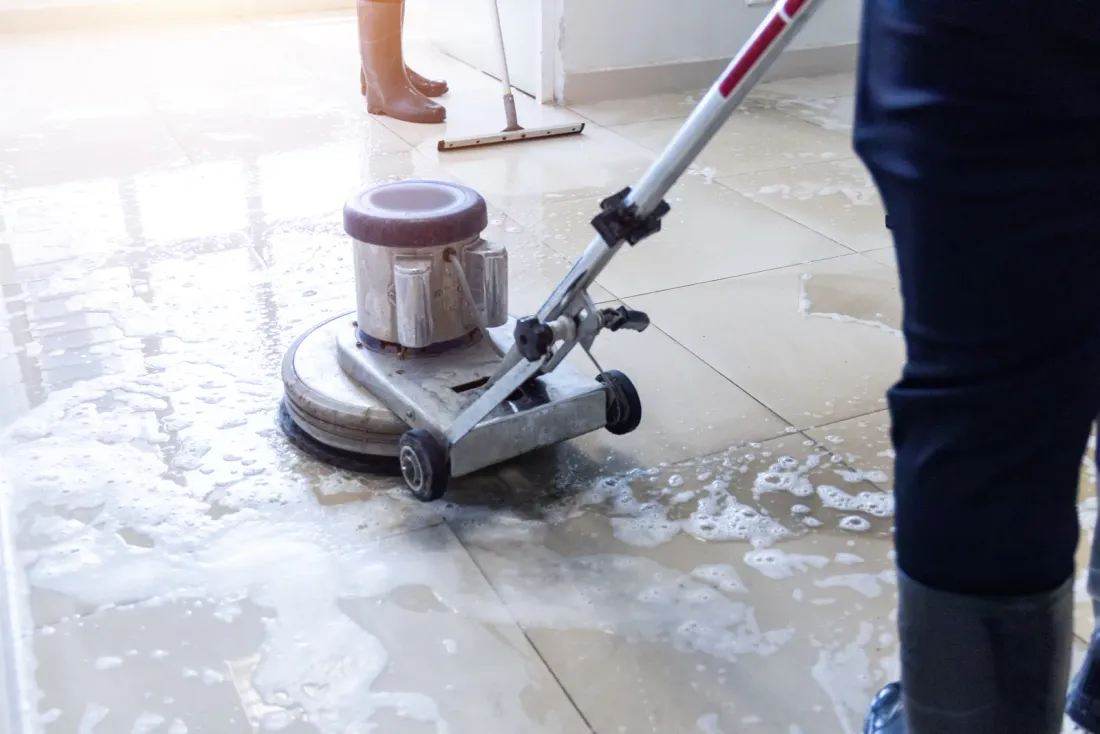 A worker running a rotary floor machine over wet, soapy tile while another person works a floor squeegee in the background, illustrating the EBS article "How Much Does Commercial Post-Construction Cleaning Cost?"