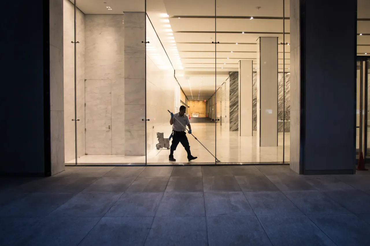 A worker wet-mopping a marble-clad lobby at night, seen from outside through a glass entrance, for the EBS guide "Deep Cleaning vs. Regular Cleaning for Commercial Spaces"