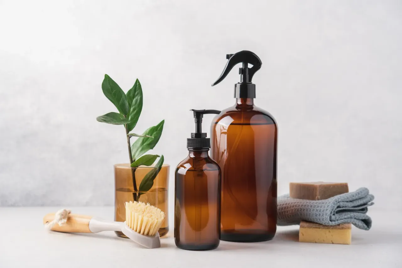 Amber glass pump and spray bottles with wooden brushes, soap bars, a folded cloth and a small plant on a pale surface, related to green cleaning services in San Bernardino County