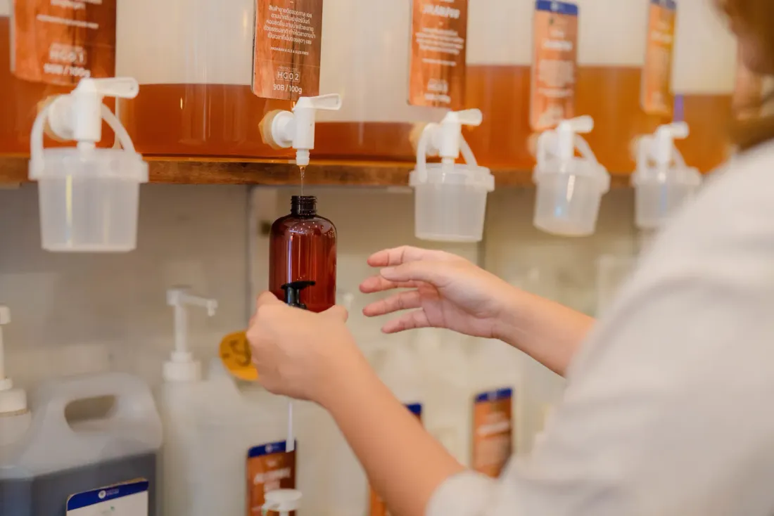 A person filling an amber bottle from a dispenser at a refill station with rows of liquid containers, related to office cleaning services in Orange, CA