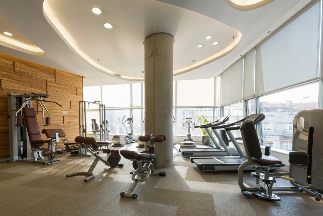 A hotel fitness room with weight machines, treadmills, a concrete column and window blinds, where a day porter keeps common areas tidy during business hours in La Jolla, CA