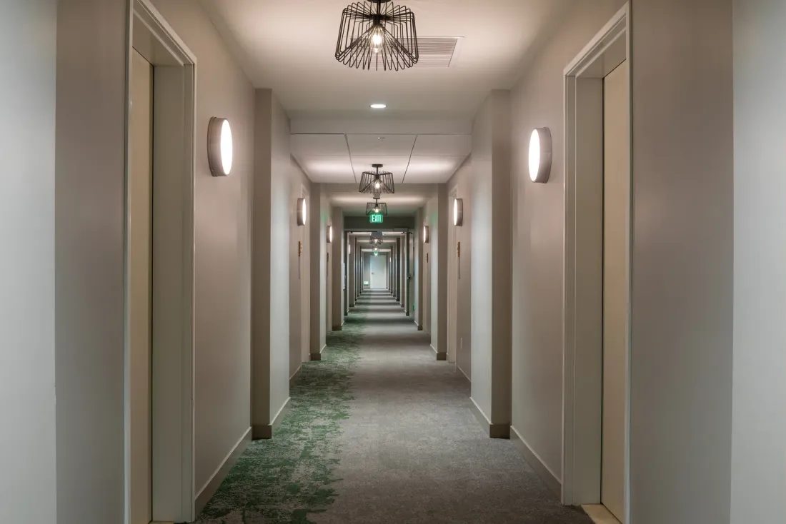 A long hotel corridor with beige walls, patterned carpet, wall sconces and wire pendant lights, a common area that day porter service keeps ready in Anaheim, CA