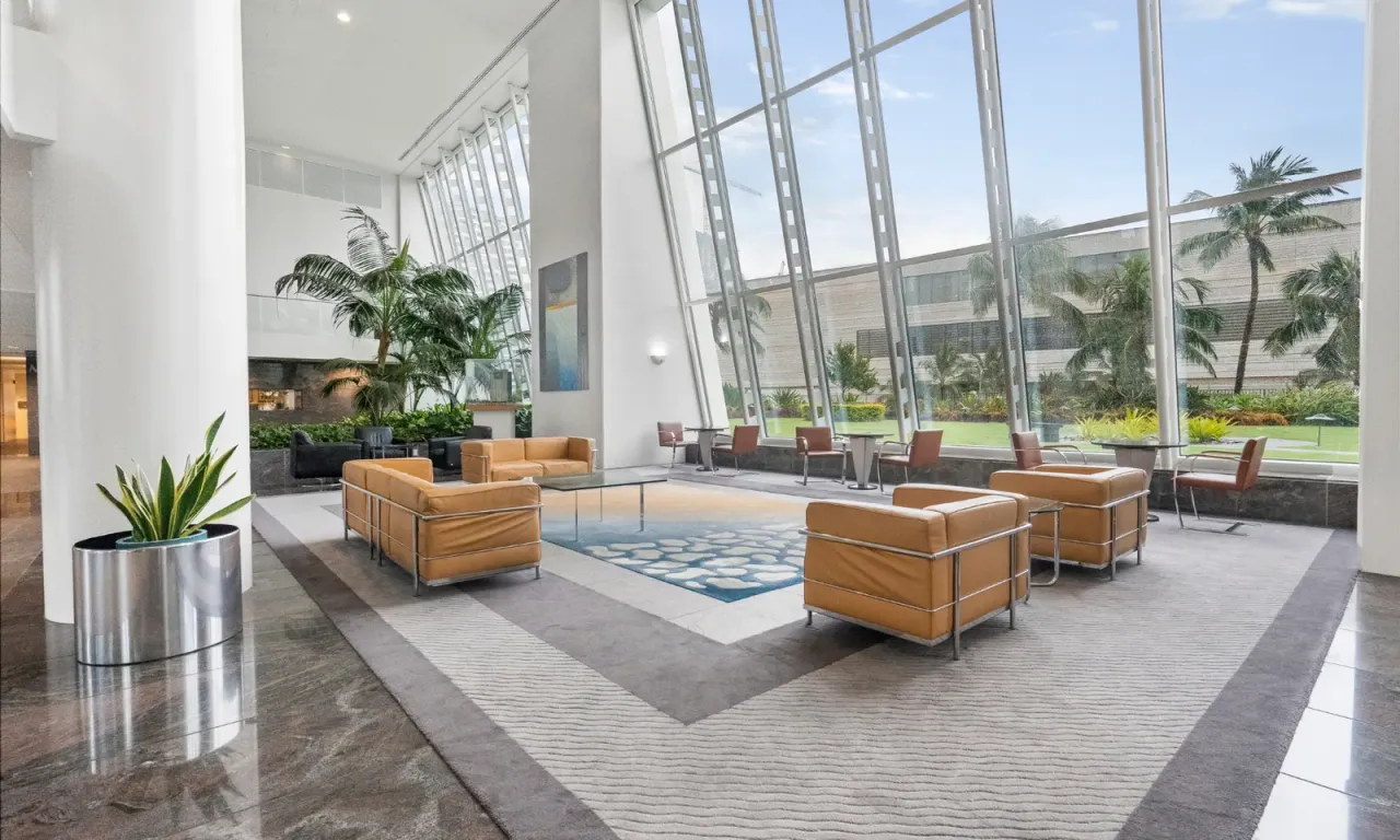 A tall, bright lobby with tan leather armchairs on a patterned rug, a plant in a steel planter and a glass wall looking out onto palm trees, an example of the facilities EBS cleans on a set schedule in Monrovia, CA