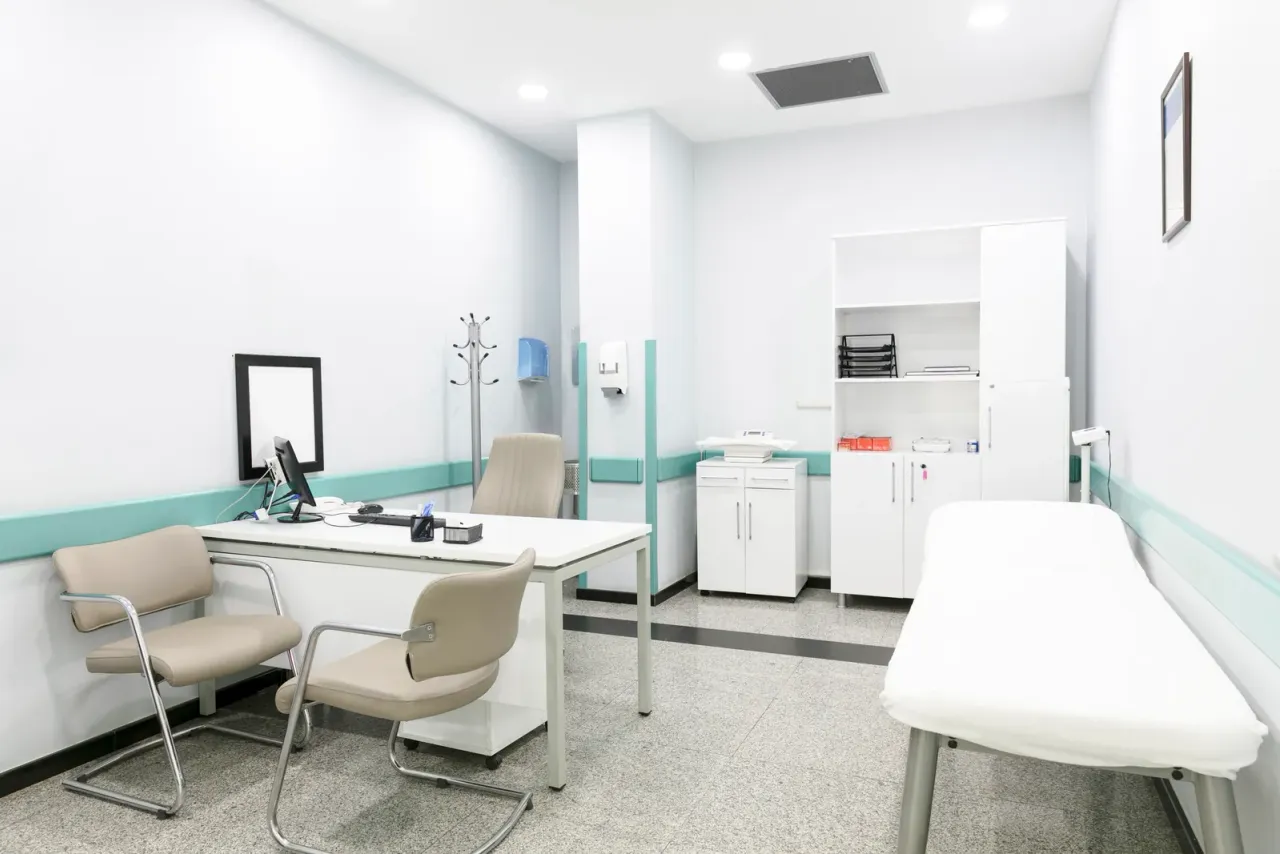 A white consultation room with a desk, two beige chairs, an examination couch and a cabinet, typical of the medical offices served by EBS in Temecula, CA