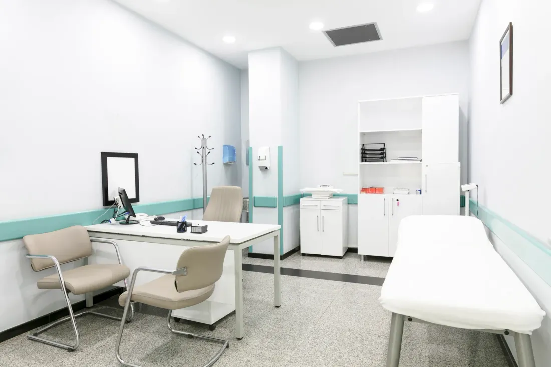 A white consultation room with a desk, two beige chairs, an examination couch and a cabinet, an example of the facilities EBS cleans on a set schedule in Inglewood, CA
