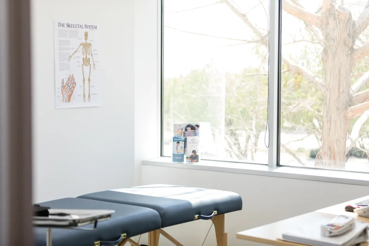 A treatment room with a padded table, a desk with papers, a skeleton chart on the wall and a large window onto trees, illustrating the EBS article "Why Do Commercial Cleaners Use Color-Coded Microfiber?"