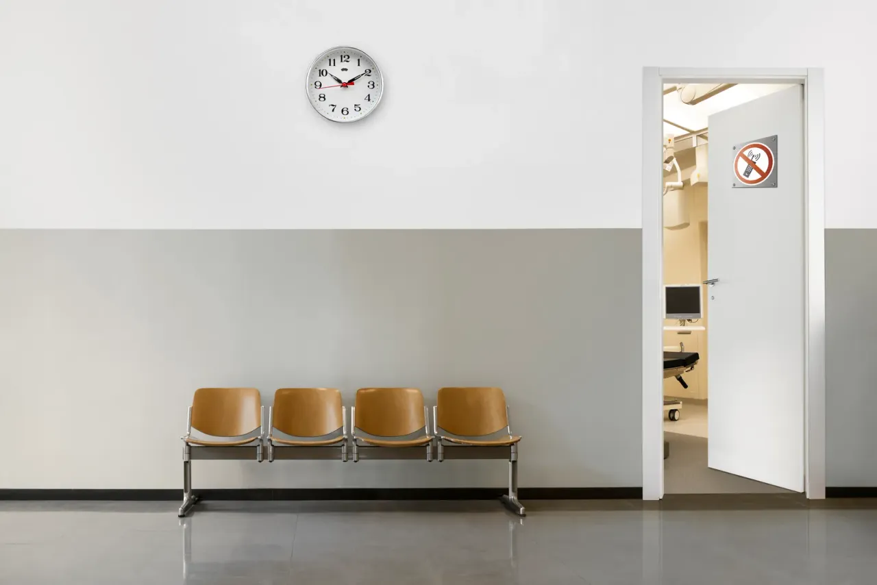A row of four tan seats against a grey and white wall, with a wall clock and an open door to a treatment room, the kind of healthcare setting EBS cleans with EPA-registered disinfectants in Riverside, CA
