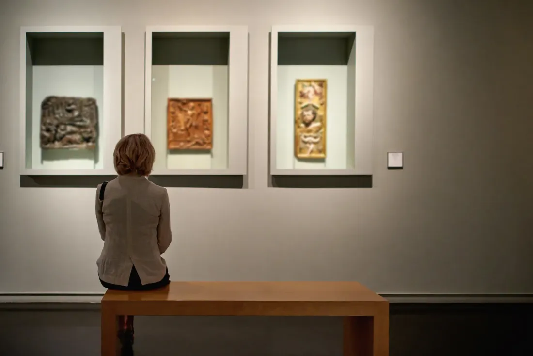 A woman seated on a wooden bench looking at three relief artefacts set into lit wall niches in a dim gallery, the kind of workspace covered by office cleaning in Miracle Mile, CA
