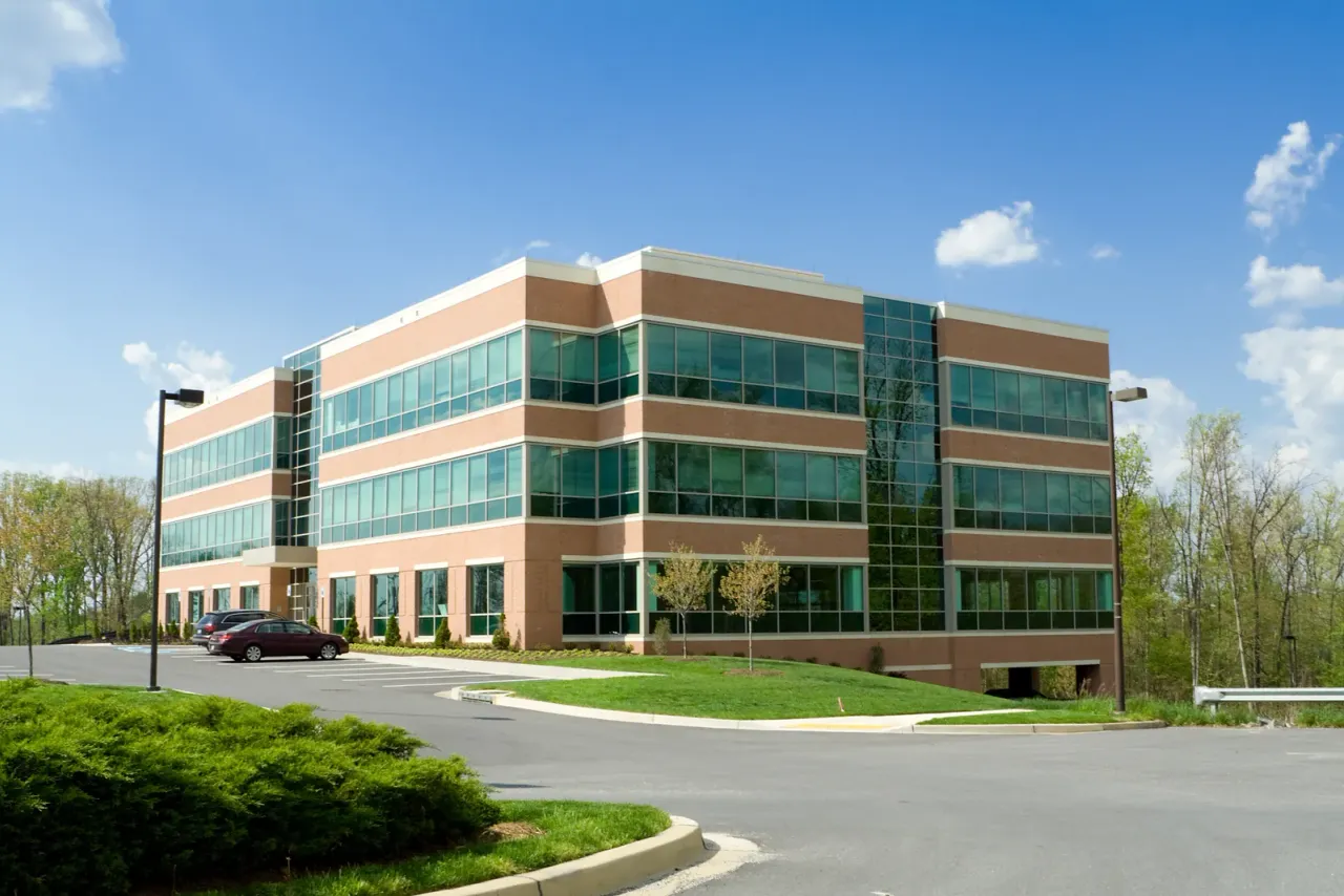 A three-storey brick office building with bands of green glass windows, a parking lot and a landscaped curb under a blue sky, like the offices EBS cleans for tenants in Studio City, CA