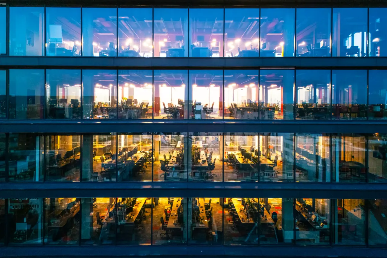 A glass office facade at dusk with lit, empty open-plan floors visible through the windows, for the EBS guide "Office Cleaning Checklist: Daily, Weekly and Monthly Tasks"