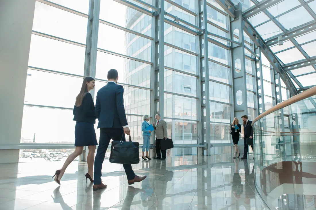 People in business clothes walking across a bright, glass-walled atrium with a polished floor, the kind of shared space day porters look after in Orange, CA