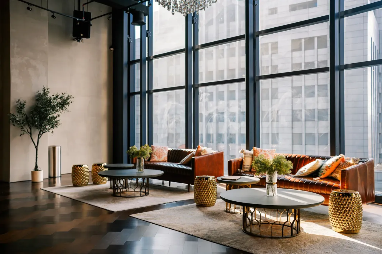 A lobby lounge with brown leather sofas, round coffee tables, potted trees and tall black-framed windows, an example of the facilities EBS cleans on a set schedule in Tustin, CA