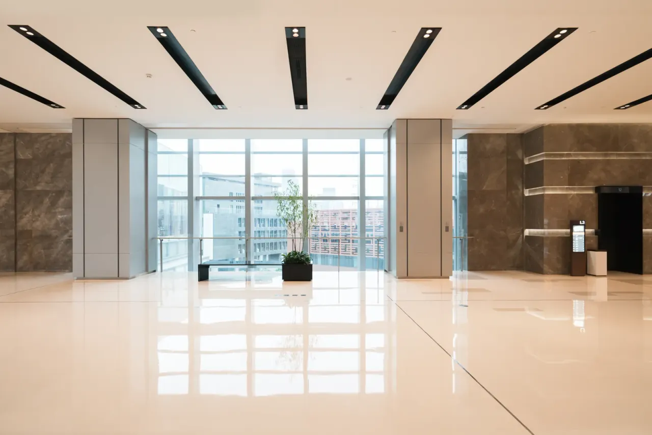 An empty lobby with polished cream floor tiles, stone-clad walls, a small potted plant and floor-to-ceiling windows, an example of the facilities EBS cleans on a set schedule in Paramount, CA