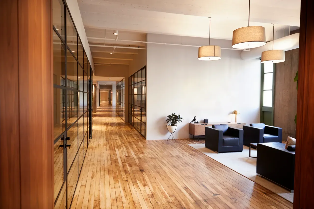 A wood-floored office with a glass-walled corridor on one side and dark armchairs around a low table under drum pendant lamps, like the offices EBS cleans for tenants in Cerritos, CA