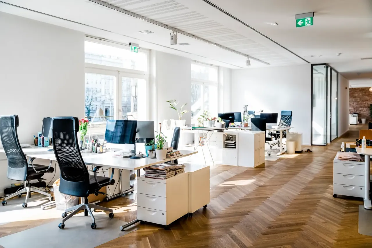An empty open-plan office with white desks, black mesh chairs, monitors and a herringbone wood floor lit by tall windows, an example of office space EBS crews clean after hours in El Segundo, CA