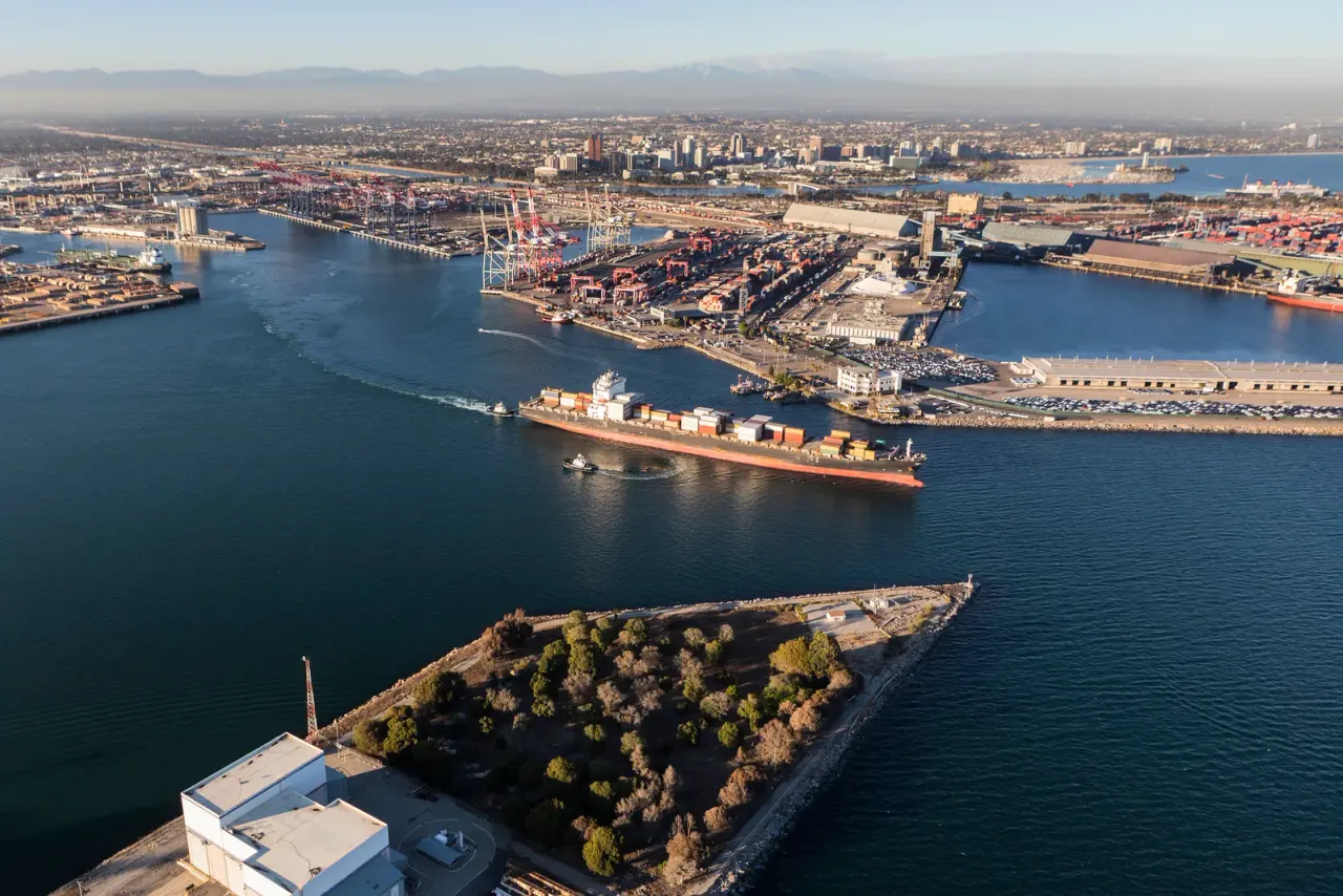 An aerial view of a container ship moving through a harbor channel past container terminals, with a city beyond, an example of the spaces EBS cleans for this sector in San Diego County