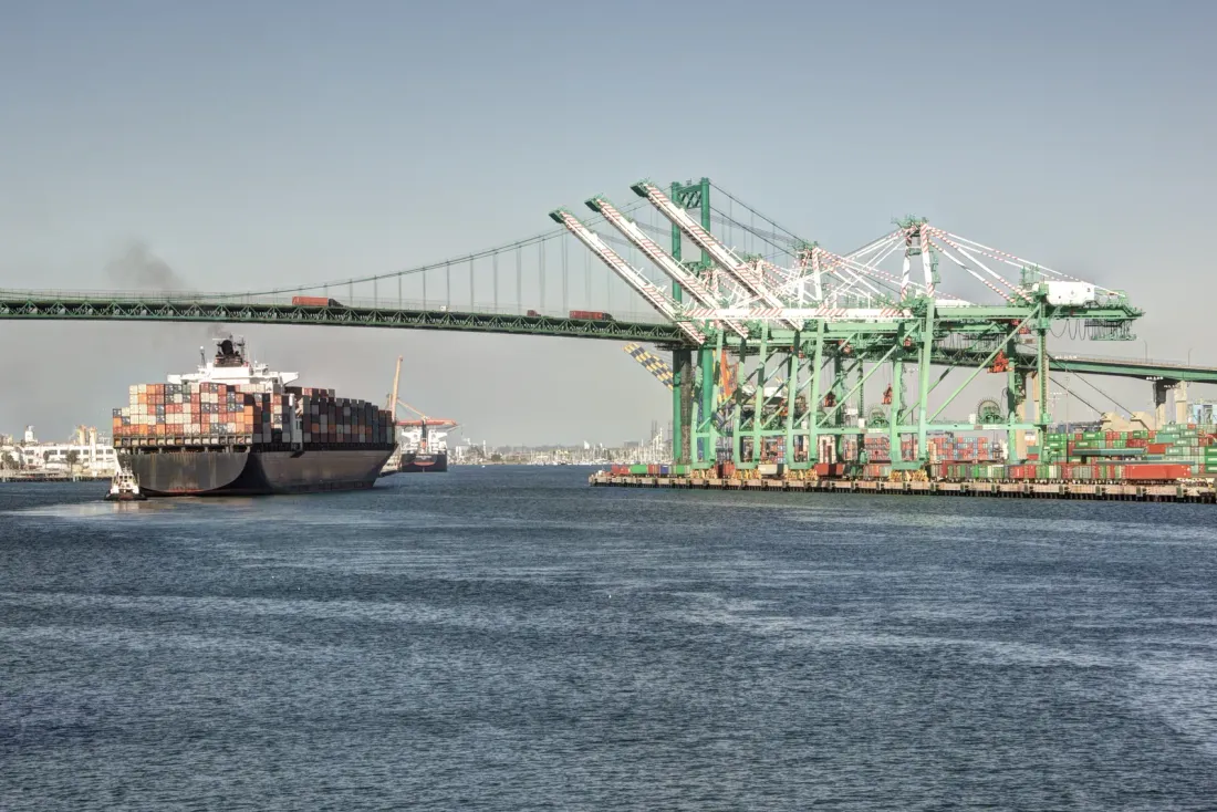Green ship-to-shore cranes at a container terminal, a loaded container ship on the water and a suspension bridge behind, the kind of space covered by warehouse cleaning services across Los Angeles County