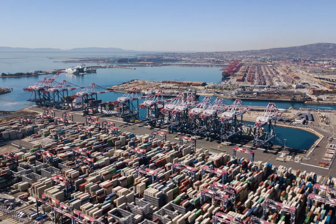 An aerial view of a container port with stacked containers, rows of gantry cranes and berths along the water under a hazy sky, typical of the business properties served by EBS in National City, CA