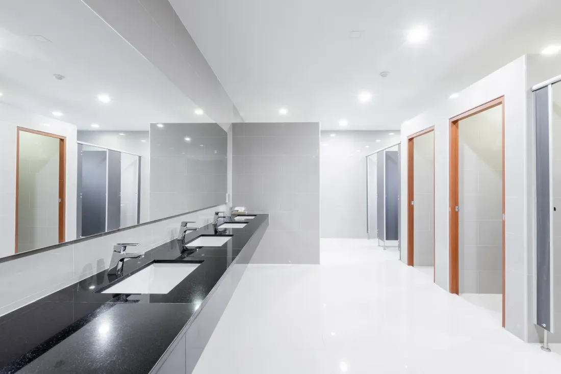 A bright restroom with undermount sinks in a black stone counter, a long mirror and a row of toilet stalls, the kind of space EBS janitorial crews maintain in Temecula, CA