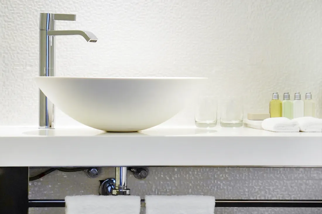 A white vessel sink with a tall chrome faucet on a white counter with folded towels and small toiletry bottles, illustrating disinfection services in Orange County
