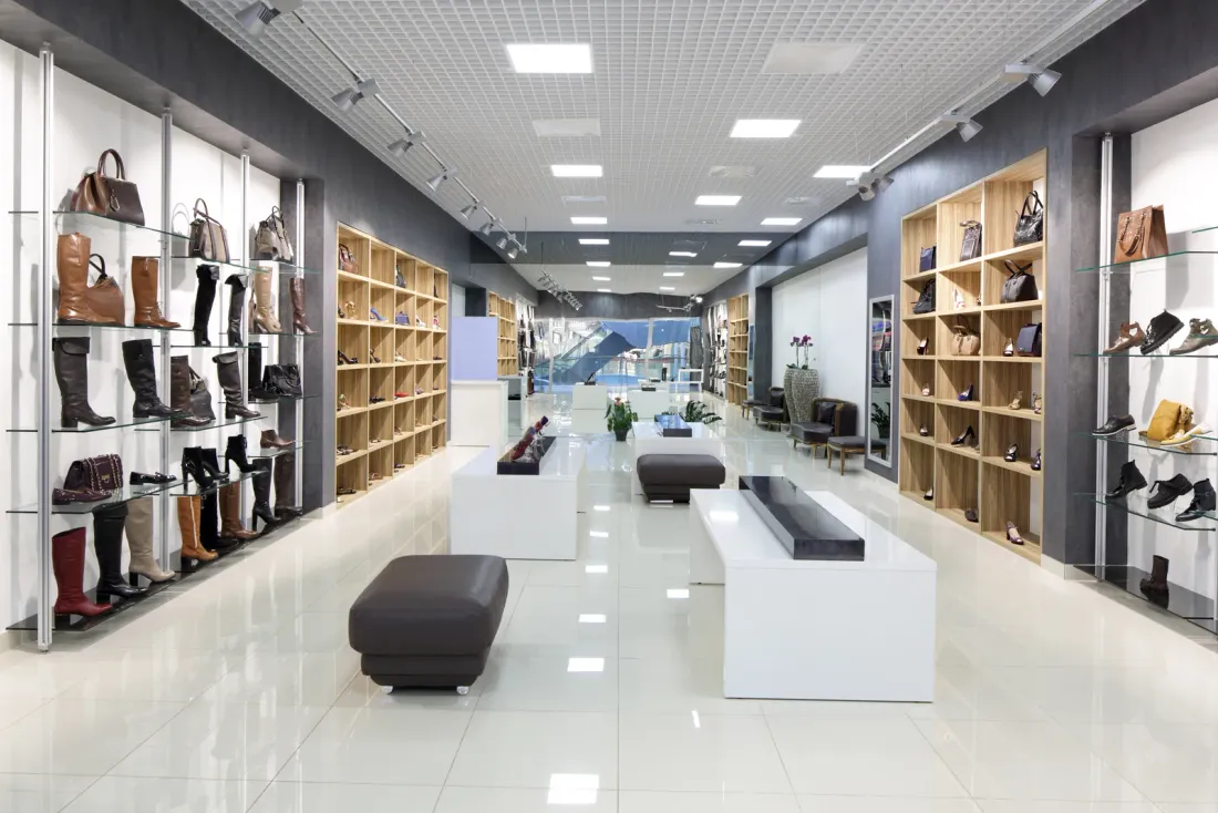 A bright shoe and handbag store with lit wall shelving, a white tile floor and low display benches, illustrating the EBS article "Exterior Window Cleaning for Commercial Buildings"