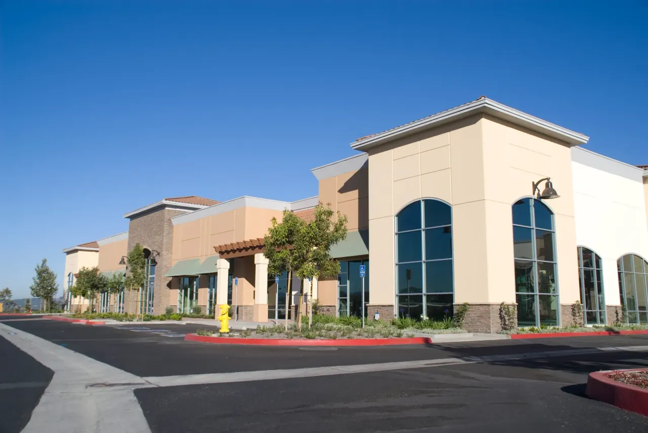 A tan shopping center with tall arched windows, young trees and an empty parking lot under a clear sky, typical of the business properties served by EBS in Montclair, CA