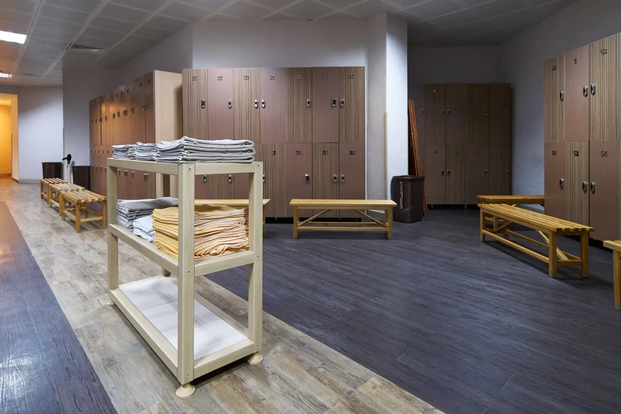 A locker room with wooden lockers, slatted benches and a cart of folded towels on a dark floor, typical of the spaces EBS cleans for this industry in Southern California