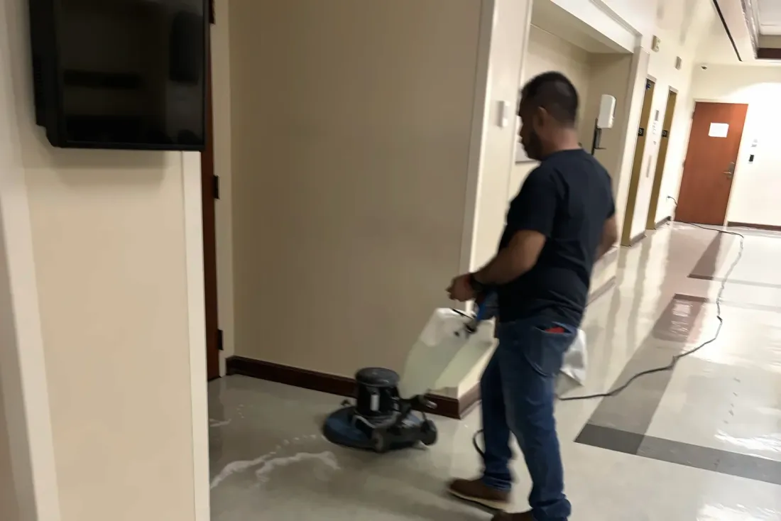 A man in a dark T-shirt running a floor buffer across a polished floor in a corridor with wooden doors, part of the EBS Maintenance team serving Carson, CA