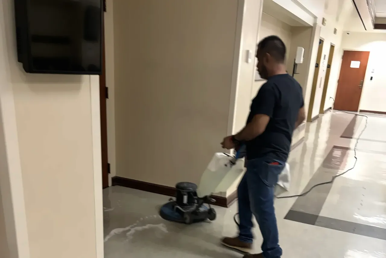 A man in a dark T-shirt running a floor buffer across a polished floor in a corridor with wooden doors, EBS staff at work, the same crews that serve Southern California