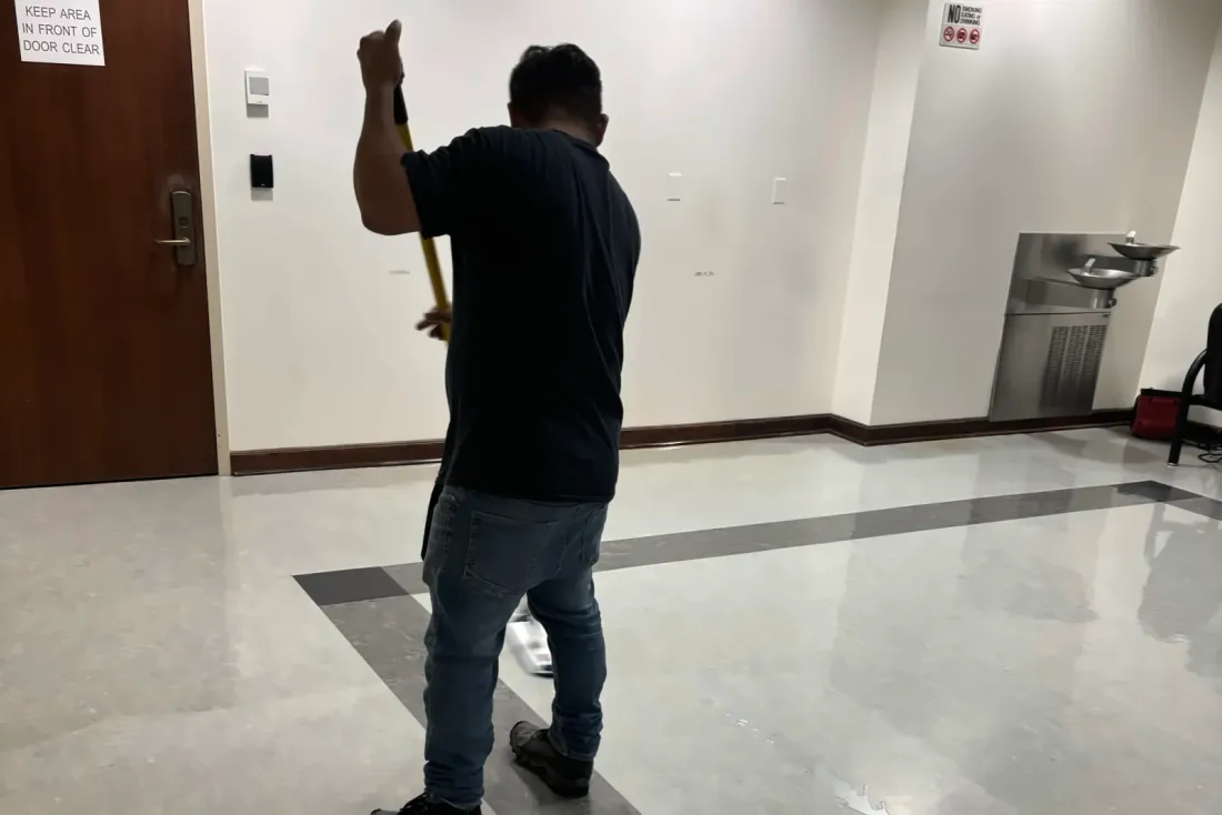 A man in a black T-shirt, seen from behind, pushing a dust mop across a polished lobby floor near a wooden door, part of the EBS Maintenance team serving Chino, CA