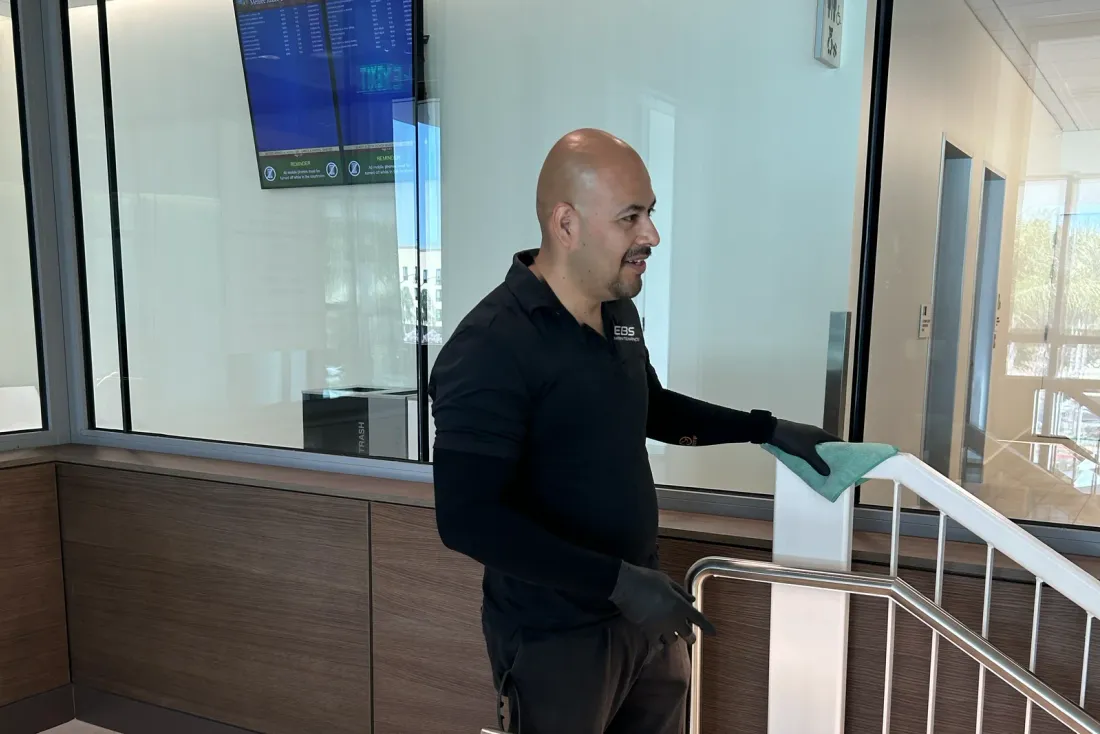 A man in a black long-sleeve shirt with the EBS logo and black gloves wiping a stair railing with a green cloth beside a glass wall, the EBS team behind cleaning programs in Murrieta, CA